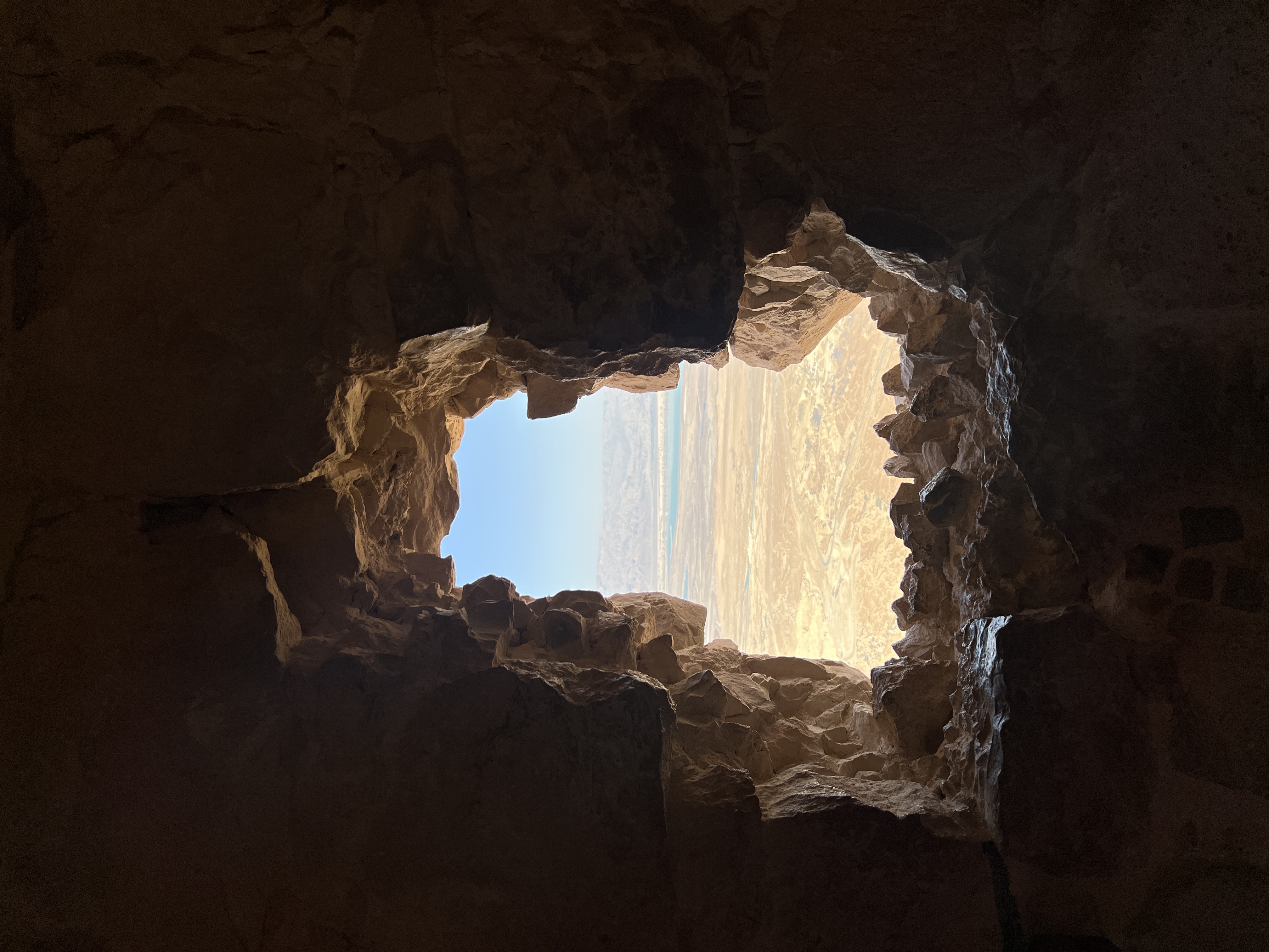 Looking through a jagged hole in ancient stone cave wall toward bright desert landscape and blue sky beyond