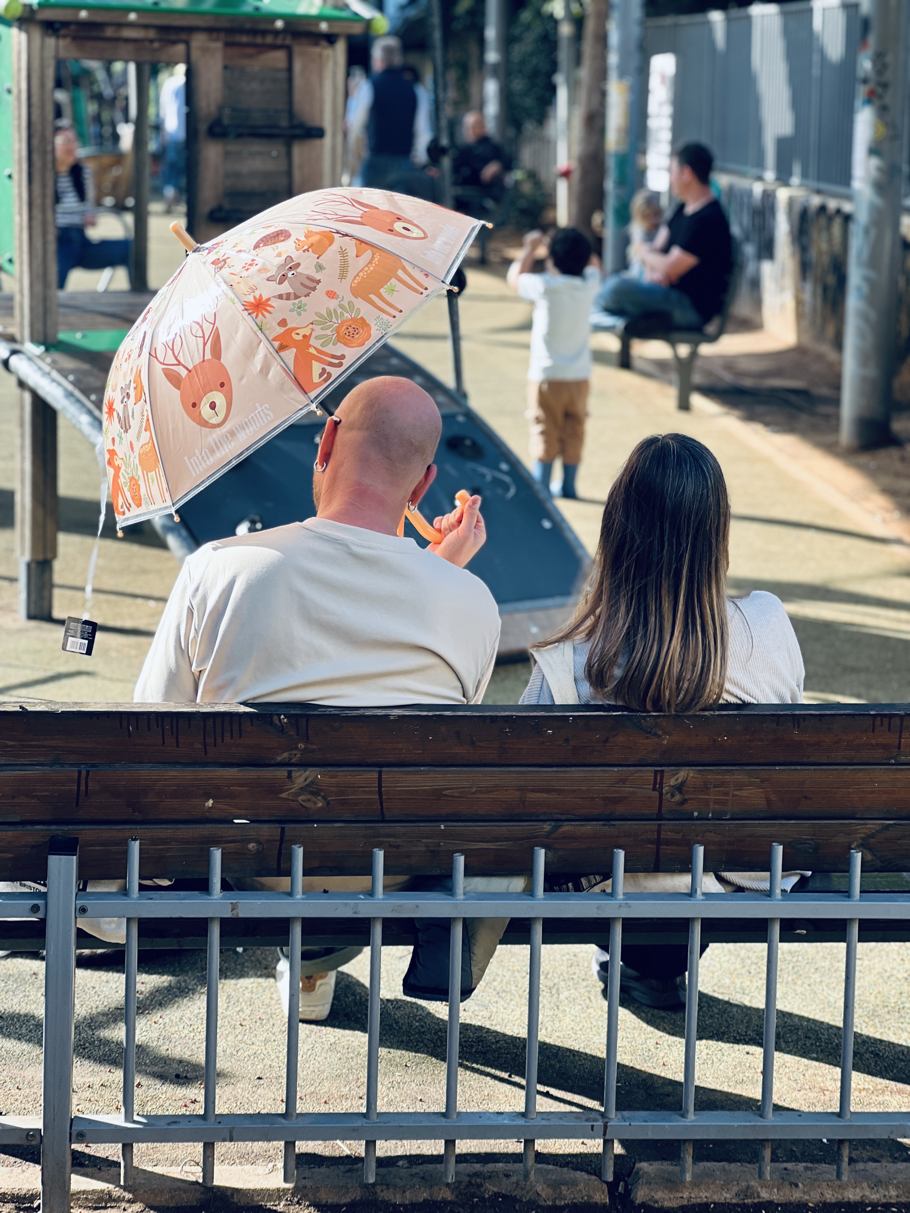 Couple sitting on bench from behind, holding colorful children's umbrella with animals, playground in background