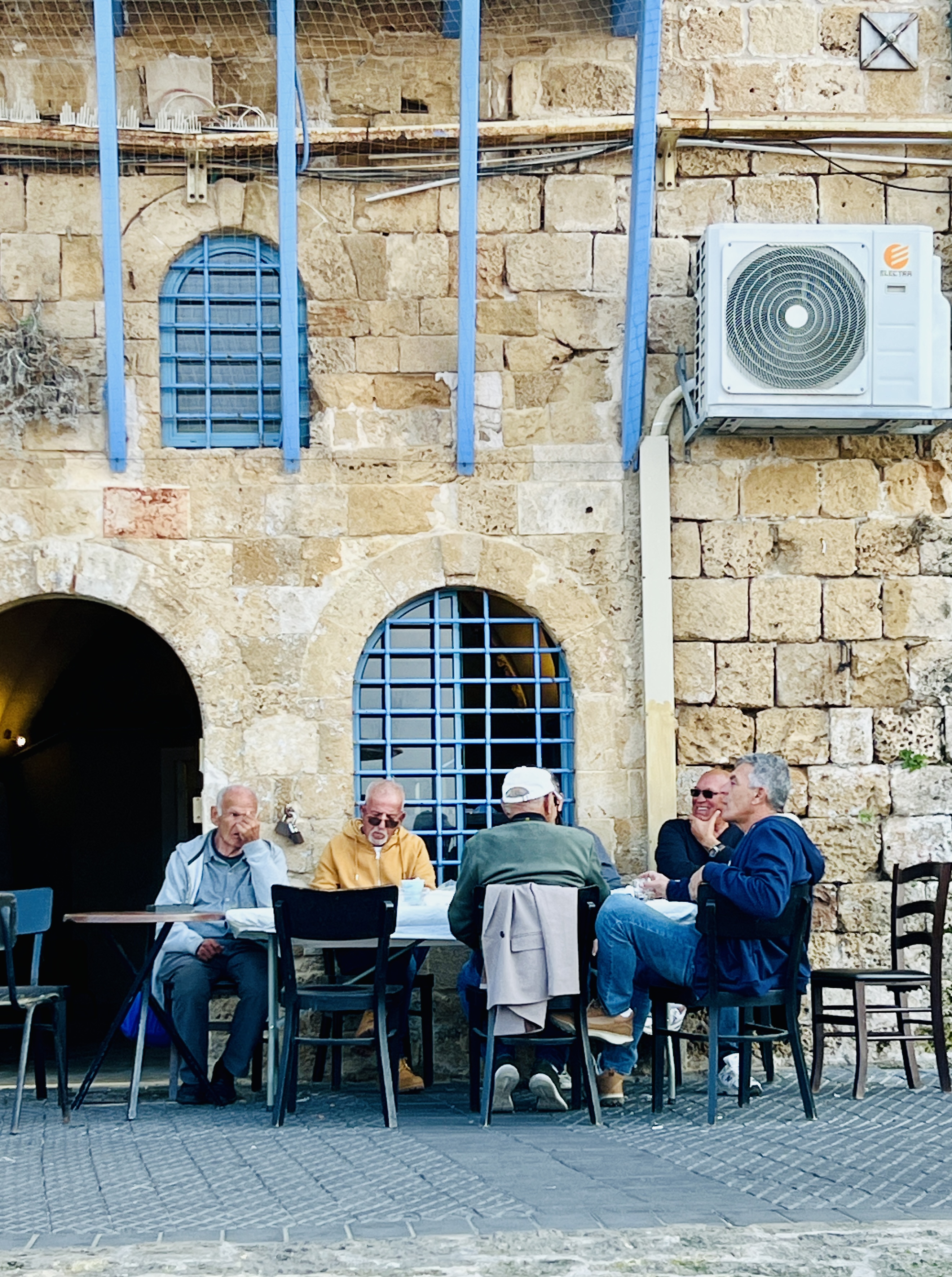 Four elderly men sitting at outdoor cafe table against ancient stone building with blue shutters in Jaffa old city