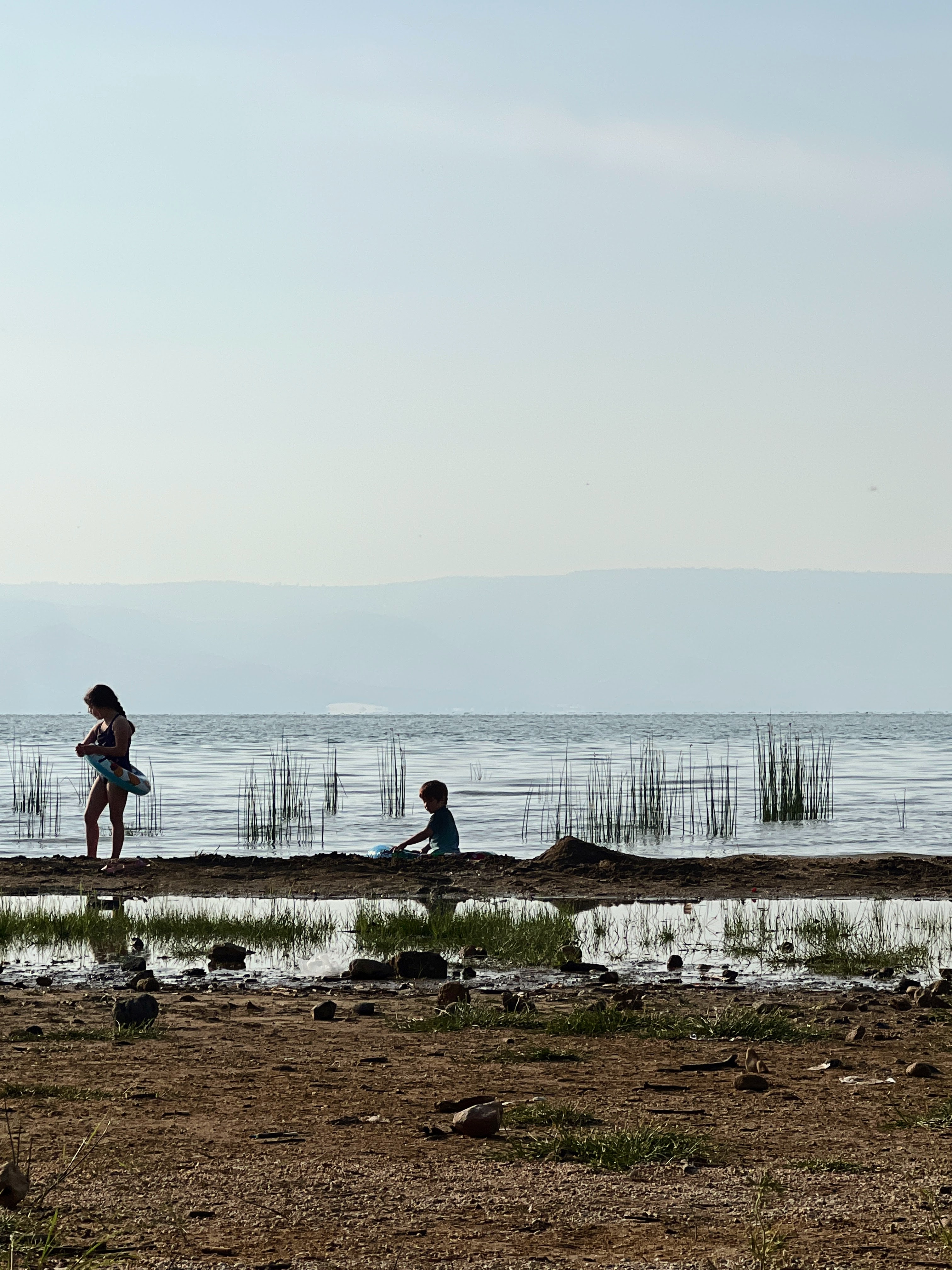 Two silhouettes of woman and child playing on the shore of the Sea of Galilee, reeds in calm water, misty mountains in background