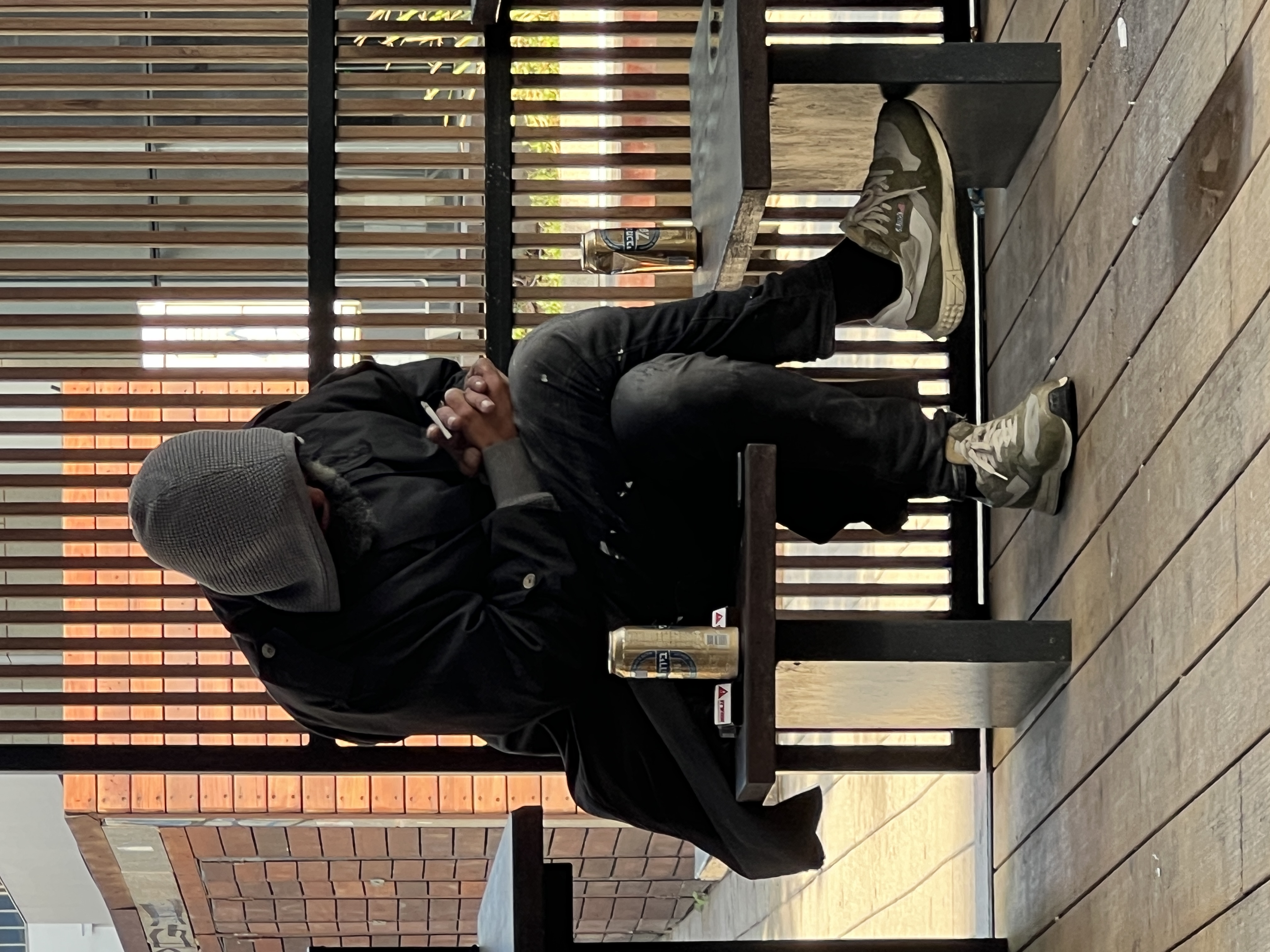 Man in dark jacket and cap sitting at cafe table smoking cigarette, wooden slat wall and warm light behind him
