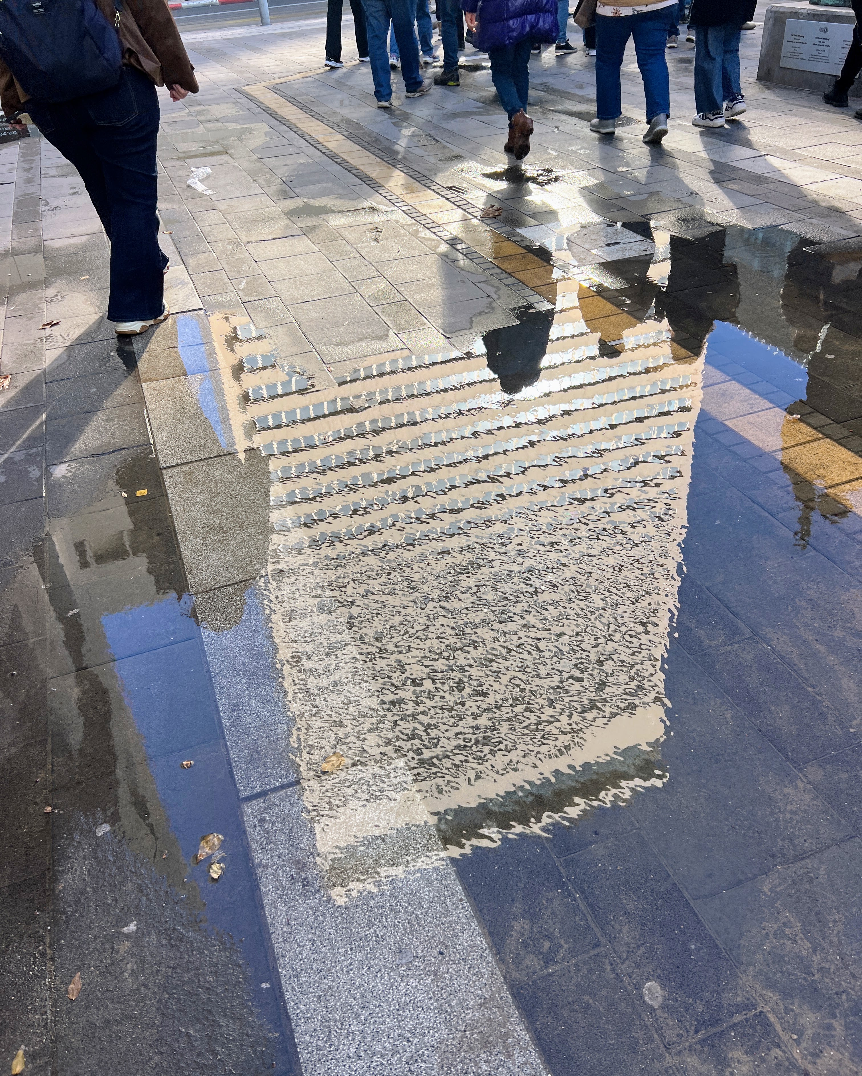 Reflection of tall building in puddle on city street with pedestrians walking in background, Tel Aviv