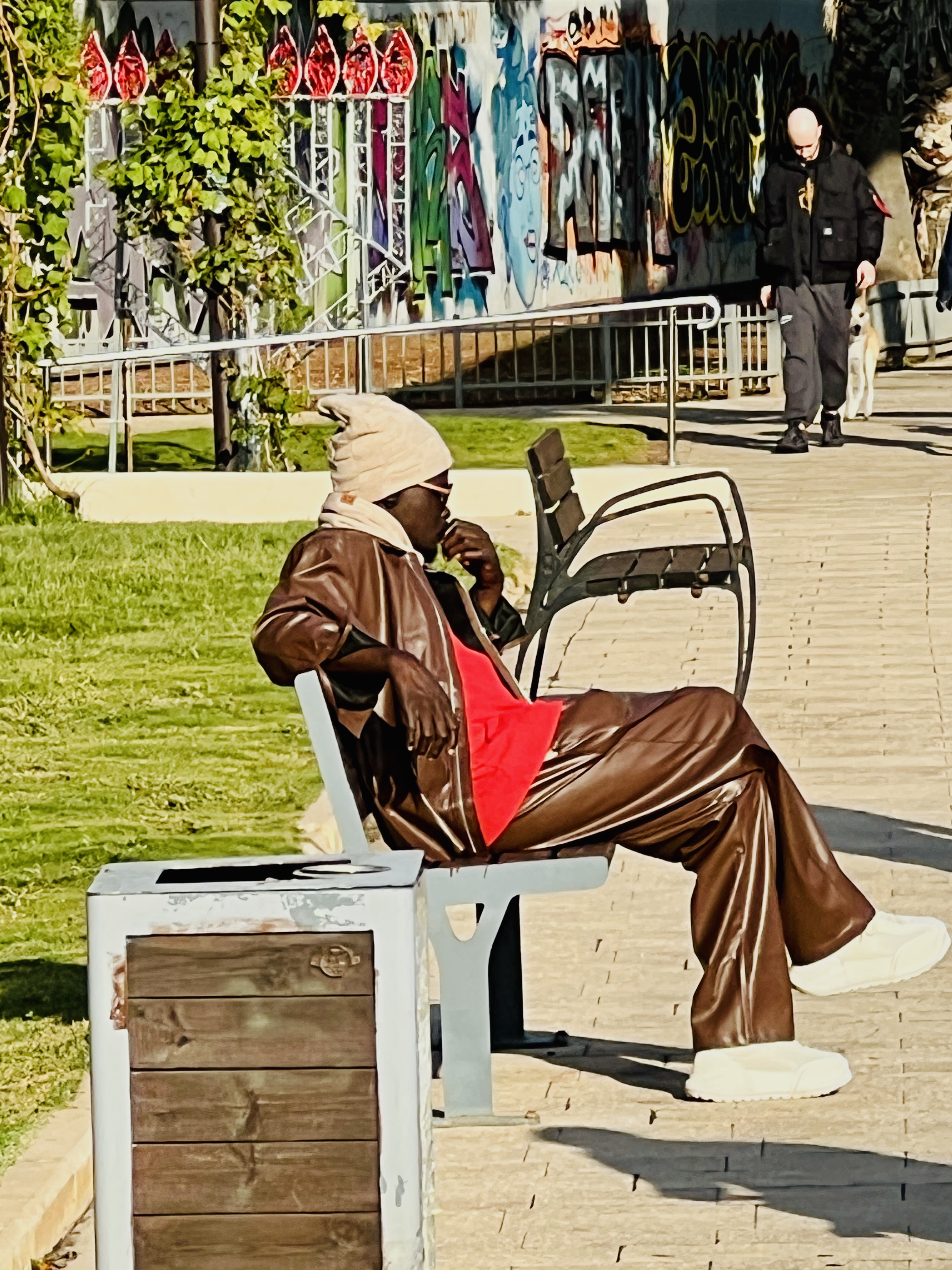 Person in brown leather coat and red skirt sitting on bench in front of colorful graffiti wall, Tel Aviv