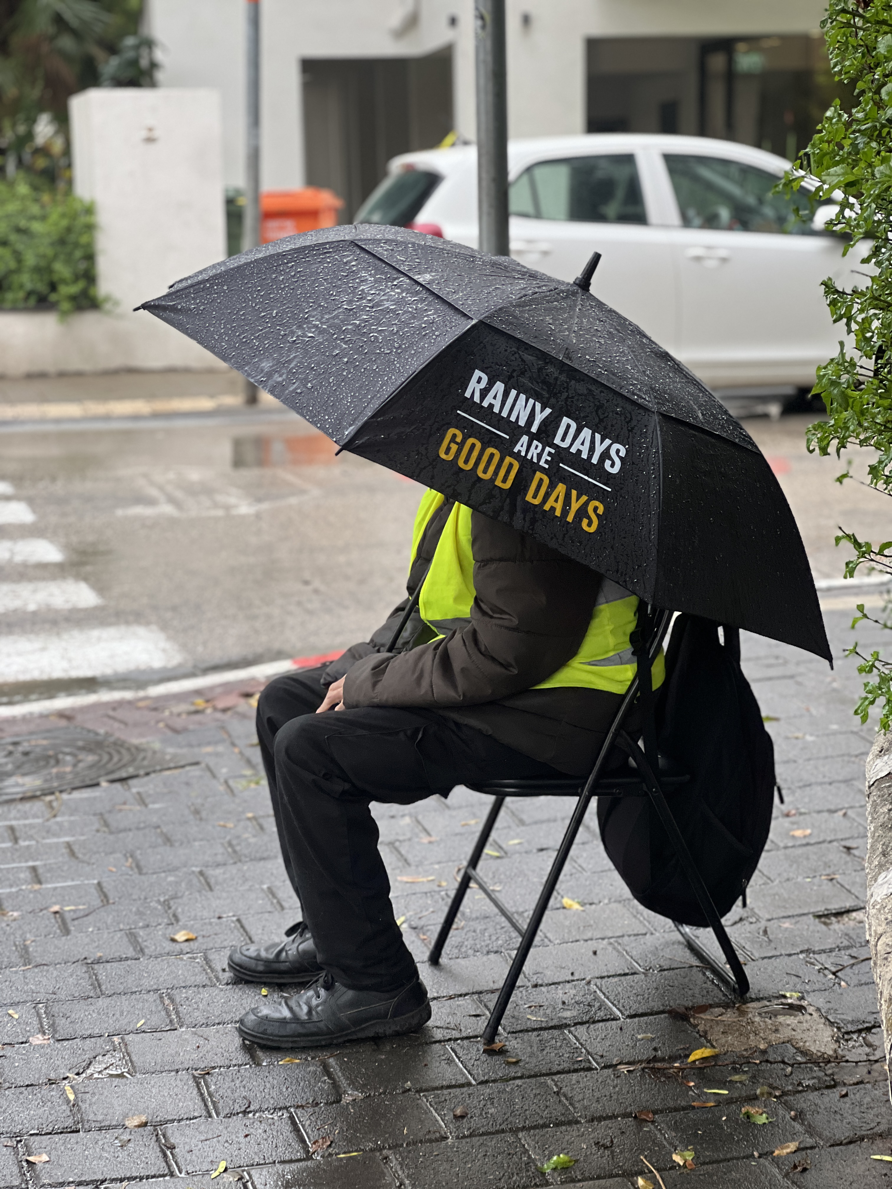 Worker in safety vest sitting on folding chair under black umbrella reading Rainy Days are Good Days on wet sidewalk